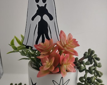 May include: A white ceramic planter with a pink abstract shape and black starburst designs, containing pink-tipped succulent foliage and a white ghost-shaped plant stake featuring a black silhouette of a cat standing inside the ghost.