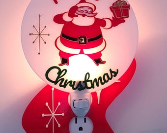 May include: A circular plug-in night light featuring a red and white Santa Claus graphic holding a gift, with the text Merry Christmas in red and green script. The base is a red, organic-shaped acrylic piece with white starburst accents.
