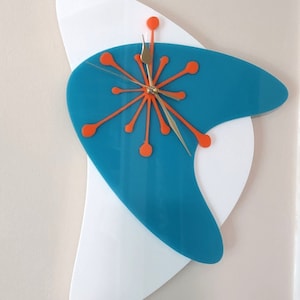 May include: A turquoise and white abstract wall clock with orange accents. The clock face features a starburst design with gold hands.