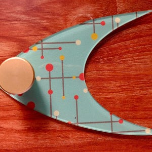 May include: A decorative, crescent-shaped object with a light blue surface. The surface features a pattern of red, yellow, and white dots connected by gray lines. A circular, bronze-colored knob is attached to the object. The item rests on a reddish-brown wooden surface.