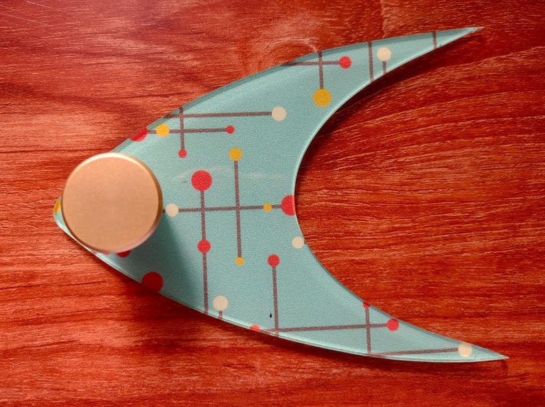 May include: A decorative, crescent-shaped object with a light blue surface. The surface features a pattern of red, yellow, and white dots connected by gray lines. A circular, bronze-colored knob is attached to the object. The item rests on a reddish-brown wooden surface.