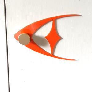 Puede incluir: Una decoración de pared en forma de pez naranja, de estilo moderno de mediados de siglo, con un pomo circular dorado. El pez tiene un recorte en forma de diamante y está montado sobre una superficie blanca.