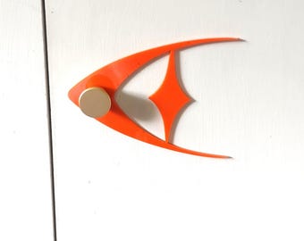 May include: An orange, mid-century modern style fish-shaped wall decor with a gold-colored circular knob. The fish has a diamond-shaped cutout and is mounted on a white surface.