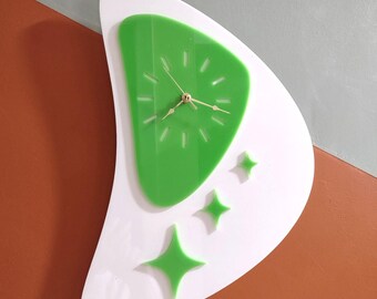 May include: A crescent-shaped wall clock featuring a white base with a green, rounded triangular center. The clock face has gold-colored hands and simple tick marks. Three green, four-pointed star shapes are arranged below the clock face.
