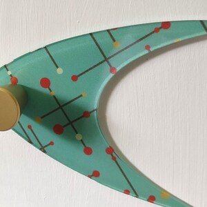 May include: A mid-century modern wall hook with a teal boomerang-shaped design. The hook features a pattern of red and yellow dots connected by black lines. It has a gold-colored cylindrical knob for hanging items.