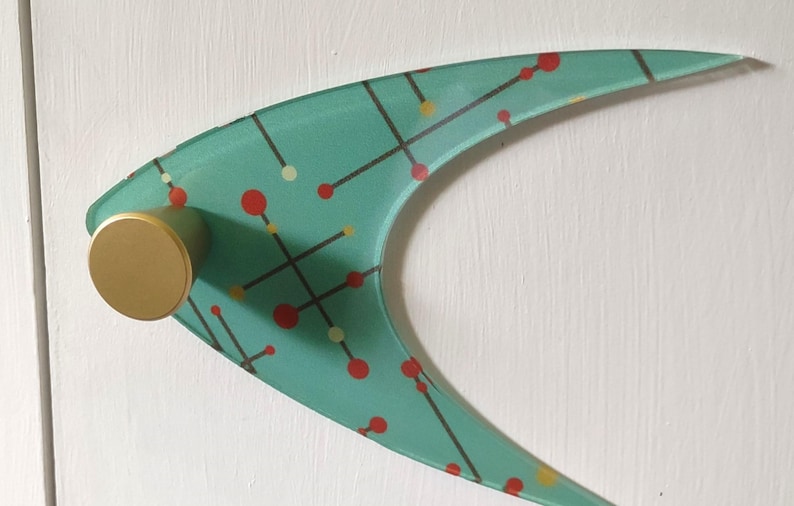 May include: A mid-century modern wall hook with a teal boomerang-shaped design. The hook features a pattern of red and yellow dots connected by black lines. It has a gold-colored cylindrical knob for hanging items.