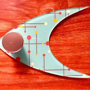 May include: A light blue, crescent-shaped decorative object with a circular, rose-colored knob. The object features a mid-century modern design with a pattern of red, yellow, and white dots connected by gray lines. The background is a reddish-brown wood grain.