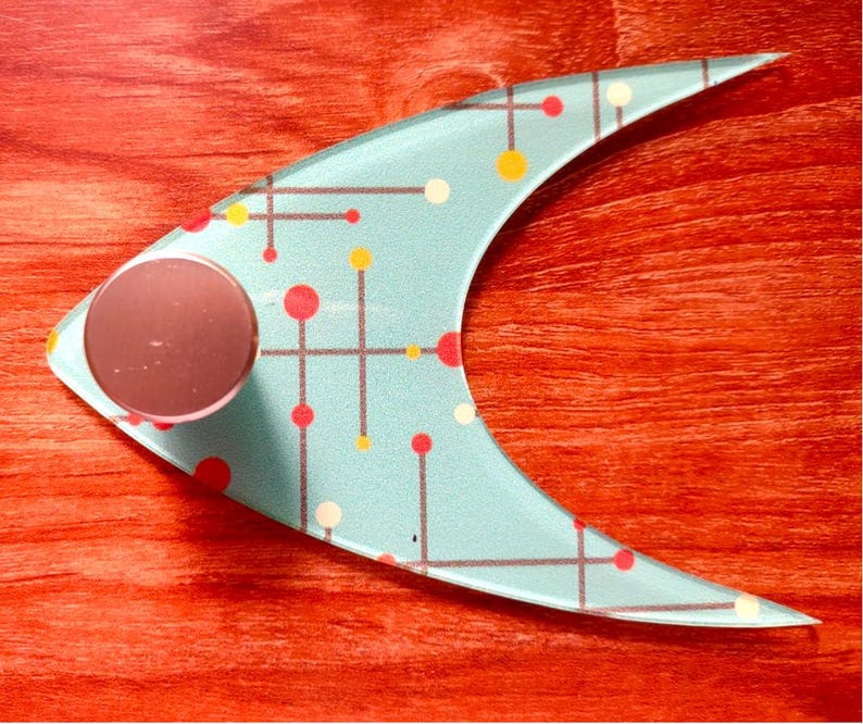 May include: A light blue, crescent-shaped decorative object with a circular, rose-colored knob. The object features a mid-century modern design with a pattern of red, yellow, and white dots connected by gray lines. The background is a reddish-brown wood grain.