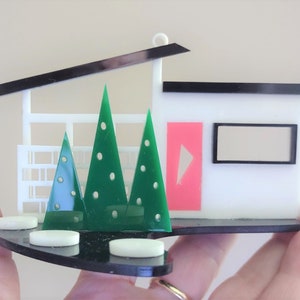 Puede incluir: Un adorno de casa moderno blanco y negro con una puerta rosa y árboles verdes. La casa tiene un techo negro y una base negra. El adorno está hecho de acrílico.