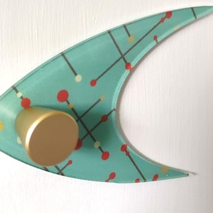 May include: A mid-century modern wall decoration. The teal-coloured, boomerang-shaped glass has a pattern of red, yellow, and white dots connected by black lines. A gold-coloured cylindrical knob is attached to the glass.
