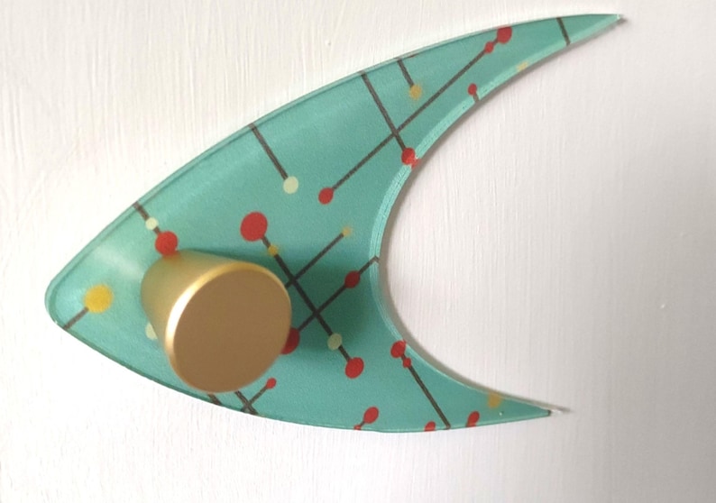 May include: A mid-century modern wall decor piece. The aqua-colored, boomerang-shaped glass has a pattern of red, yellow, and white dots connected by black lines. A gold-colored cylindrical knob is attached to the glass.