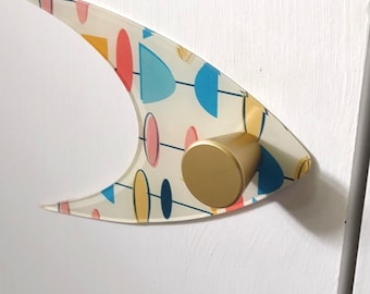 May include: A boomerang-shaped wall hook featuring a cream background with a colorful pattern of red, yellow, and blue geometric shapes, including circles and semi-circles, connected by thin lines, finished with a solid gold-toned cylindrical knob.