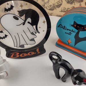May include: A set of three Halloween-themed night lights. The first is a round, black light with a white ghost and a black cat. The second is a set of four coasters with a turquoise background and a black cat silhouette. The third is a black cat figurine. The coasters have the text "Atomic cat" on them. The round light has the text "Boo!" on it.