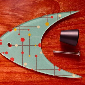 May include: A mid-century modern style fish-shaped glass cabinet pull with a teal background and a pattern of red, yellow, and white dots and lines. Includes a black handle and a silver screw. The items are on a reddish-brown wooden surface.
