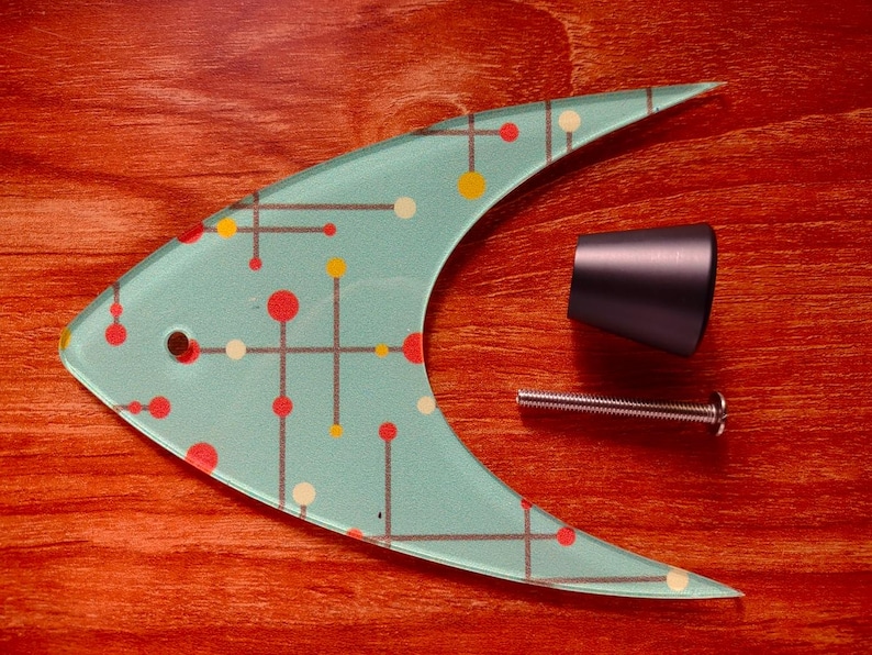 May include: A mid-century modern style fish-shaped glass cabinet pull with a teal background and a pattern of red, yellow, and white dots and lines. Includes a black handle and a silver screw. The items are on a reddish-brown wooden surface.