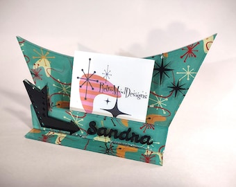 May include: A teal business card holder featuring a mid-century atomic starburst pattern in red, yellow, and black. The holder includes a black arrow accent on the left and the name Sandra in black script lettering on the front base.