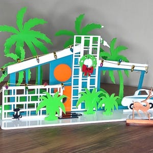 May include: A colorful, mid-century modern Christmas decoration featuring a house with a teal and orange facade, palm trees, and a small car. The house has a white fence and a wreath with a red bow. A small deer is also included.