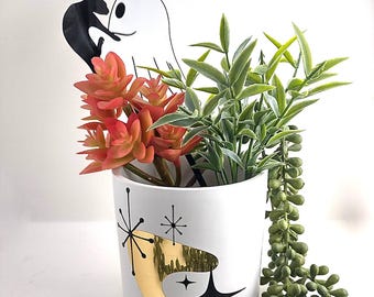 May include: A white cylindrical planter featuring a gold boomerang shape and black starburst patterns. Inside are artificial green and orange succulents. A black cat silhouette and a white ghost outline are inserted into the planter.