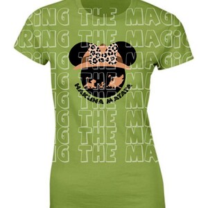 Op de afbeelding: Groen T-shirt met een witte omtrek van de zin "Bring the Magic" die meerdere keren wordt herhaald. In het midden van het shirt staat een zwart silhouet van Mickey Mouse met een safarihoed met een luipaardprint strik en een bruine safarihoed. Het silhouet bevat de zin "Hakuna Matata" in zwarte tekst.