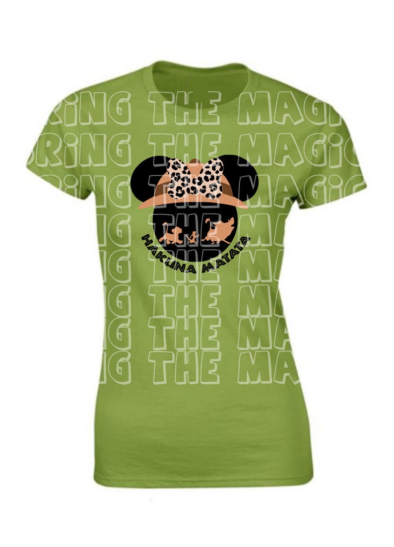 Op de afbeelding: Groen T-shirt met een witte omtrek van de zin "Bring the Magic" die meerdere keren wordt herhaald. In het midden van het shirt staat een zwart silhouet van Mickey Mouse met een safarihoed met een luipaardprint strik en een bruine safarihoed. Het silhouet bevat de zin "Hakuna Matata" in zwarte tekst.