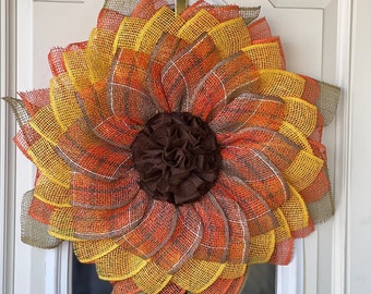 May include: A brown, orange, and yellow sunflower wreath made of burlap mesh. The wreath is hanging on a white door.
