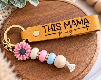 Può includere: Un portachiavi in pelle giallo senape con il testo nero THIS MAMA Prays e un simbolo a croce. Include un anello in metallo dorato, una perlina a forma di fiore rosa e una serie di perline rotonde bianche, rosa, azzurre e in legno naturale.