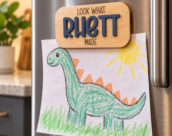 Può includere: Un magnete rettangolare in legno con angoli arrotondati che riporta la scritta "LOOK WHAT RHETT MADE" in nero, con il nome "RHETT" in lettere blu scuro in rilievo. Sotto è attaccato il disegno di un bambino con un dinosauro verde e un sole.