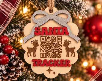 Può includere: Un ornamento in legno chiaro con bordo smerlato, caratterizzato da un fiocco glitterato argento in alto. Il testo SANTA TRACKER è in rosso, con un codice QR centrale affiancato da due illustrazioni di renne marroni e due piccole stelle.