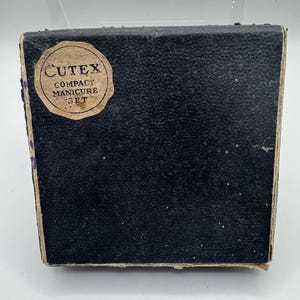 May include: A vintage black Cutex Compact Manicure Set box. The box has a textured surface and a circular label with the brand name and product description. The edges of the box show signs of wear and tear, indicating its age.
