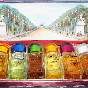 Op de afbeelding: Een set van zes miniatuur parfumflesjes in een rode en gouden doos. De flesjes zijn gelabeld met de namen "Golden Gulls", "Mada Louberry", "August Moon", "Jaffa Louis", "Zorba" en "From Seed".