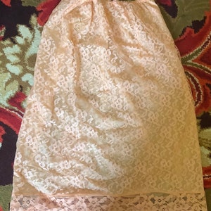 May include: A peach-colored lace slip skirt with an elastic waistband. The skirt is made of a delicate floral lace fabric and has a scalloped lace trim at the hemline. The slip skirt is designed to be worn under a dress or skirt.
