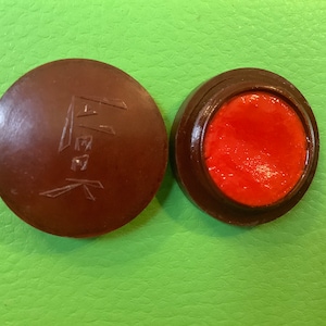 Puede incluir: Un pequeño recipiente redondo marrón con una tapa roja. La tapa tiene la palabra "真味" escrita en caracteres chinos. El recipiente está abierto y muestra una sustancia roja y cerosa en su interior.