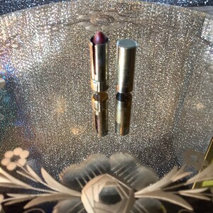 May include: Two gold lipstick tubes with a red lipstick bullet inside. The tubes are sitting on a sparkly, silver surface.