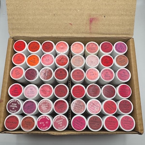 May include: A box containing many lipstick testers in shades of pink, red, and mauve. The lipsticks are in small, white containers with colour names and numbers on the lids. The box is open, showing the organised arrangement of the testers.