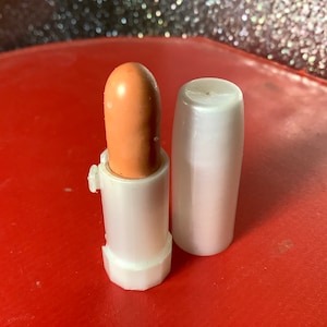 May include: A white lipstick tube with a light brown lipstick inside. The lipstick is in a twist-up tube and the cap is off.