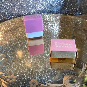 May include: Two vintage stick makeup refills. One is a stack of three blocks, with a pink, white, and lavender top. The other is a pink box with the words "Stick Makeup REFILL" in white.