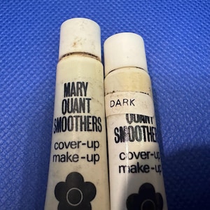 Könnte beinhalten: Zwei weiße Quetschtuben mit Mary Quant Smoothers Abdeck-Make-up. Die linke Tube ist größer, die rechte ist kleiner und mit einem Etikett mit der Aufschrift DARK versehen. Beide haben schwarzen Text und ein schwarzes Blumenlogo im unteren Bereich.