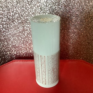 Könnte beinhalten: Weiße zylindrische Flasche mit einem goldenen und weißen Muster. Die Flasche hat einen weißen Verschluss mit einem goldenen Design.