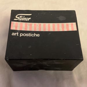 Op de afbeelding: Een zwarte doos met het woord "Steiner" in witte letters bovenaan en een roze en wit gestreepte band in het midden. De woorden "art postiche" zijn eronder in het zwart gedrukt.