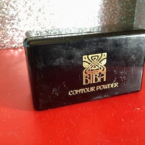 May include: A black compact with gold lettering that reads "BIBA CONTOUR POWDER".