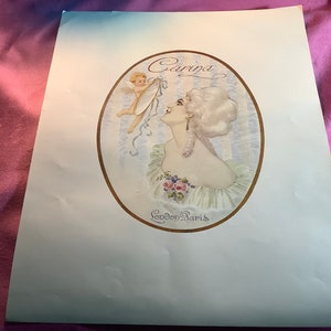 Puede incluir: Un anuncio vintage de Carina, una marca de perfume, con una mujer con una peluca blanca y un querubín sosteniendo una cinta. El texto "Carina London Paris" está escrito en un óvalo dorado.