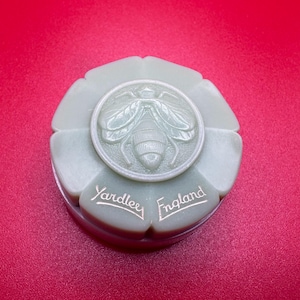 May include: A pale green, flower-shaped compact with a raised bee design in the center. The words "Yardley England" are inscribed in silver script. The compact is set against a red background.