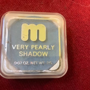 Könnte beinhalten: Ein kleiner, runder, blauer Lidschatten-Kompakt mit einem goldenen 'm'-Logo und dem Text 'Very Pearly Shadow 0.07 oz. Net Wt. 2g.'