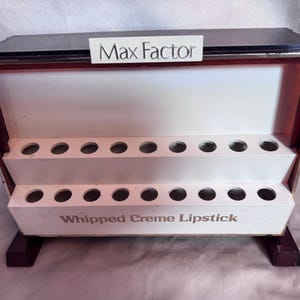 Può includere: Un espositore rettangolare Max Factor per Whipped Creme Lipstick con due ripiani bianchi a gradini dotati di fori circolari per l'alloggiamento dei prodotti. L'unità presenta una cornice marrone scuro, un'intestazione bianca con testo nero e due piedini.