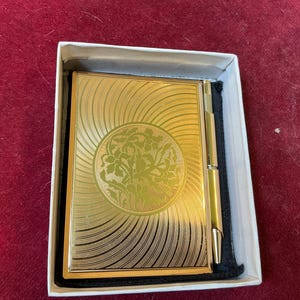 May include: A gold-toned compact mirror with a floral design in a white box. The compact has a sunburst pattern radiating from a central floral medallion. The mirror is rectangular and has a gold-colored frame and hinge.
