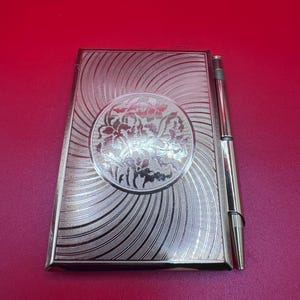 May include: A silver-toned compact with a floral design in a circular frame. The compact has a spiral pattern radiating from the center. A matching silver pen is attached to the side. The items are likely vintage.