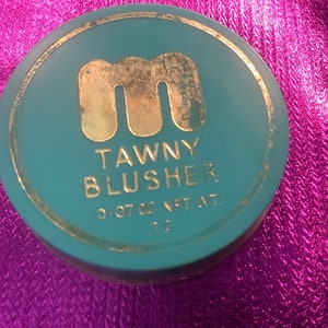 Könnte beinhalten: Eine runde, türkisfarbene Kompaktdose mit goldfarbenem Rand und einem goldenen Buchstaben "M" in der Mitte. Der Text "Tawny Blusher 0.07 oz Net Wt." ist in Gold auf der Dose aufgedruckt.