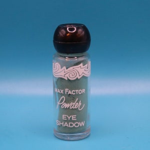 Può includere: Un piccolo barattolo di vetro di ombretto in polvere Max Factor. Il barattolo è verde e ha un tappo nero. L'etichetta dice "Max Factor Powder Eye Shadow" in lettere bianche.
