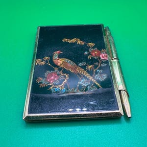 May include: A vintage black compact with a gold frame and a matching pen. The compact features a colorful illustration of a pheasant perched on a branch with flowers. The pen is gold-toned and attached to the side.