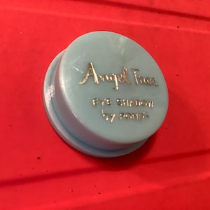 Può includere: Un contenitore rotondo di colore azzurro chiaro con un coperchio dorato. Il contenitore ha la scritta "Angel Face Eye Shadow by Pond's" stampata su di esso.